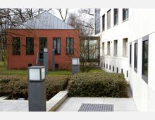 Roter, moderner Flachbau mit großen Fenstern und einem grauen Metalldach, umgeben von einer Grünanlage und kahlen Bäumen; im Vordergrund Hecken, gepflasterte Wege und quadratische Laternen. Rechts ein angrenzendes, weißes Gebäude mit rechteckigen Fenstern.
