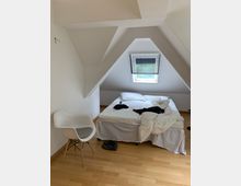Ein schlichtes Schlafzimmer in einer Dachgeschosswohnung mit weiß gestrichenen Wänden und einer Schräge, die ein kleines Fenster mit geschlossener Jalousie umfasst. In der Mitte steht ein Doppelbett mit unordentlicher weißer Bettwäsche, daneben befinden sich ein weißer Stuhl und einige Schuhe auf dem Holzboden.