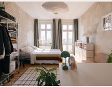 Schlafzimmer mit hellen Wänden in einem leicht rustikalen Look, einem Doppelbett mit farbiger Decke, einem offenen Metallregal für Kleidung und einem weißen Sideboard. Große Fenster mit grünen Vorhängen lassen viel Tageslicht herein, und verschiedene Dekorationsgegenstände wie Lampen und Pflanzen ergänzen den Raum.