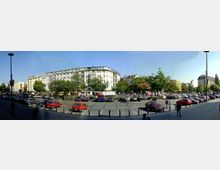 Großer Platz in einer Stadtumgebung mit parkenden Autos und Bäumen, die in der Mitte des Platzes angeordnet sind. Im Hintergrund sind mehrstöckige Wohngebäude im Stil der Jahrhundertwende sowie modernere Gebäude zu sehen.