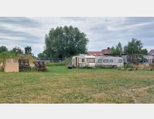 Freifläche mit Gras und zwei alten Wohnwagen, umgeben von Bäumen und vereinzelten Gebäuden im Hintergrund. Links im Bild liegen gestapelte Holzpaletten, während rechts verschiedene Gegenstände und eine einfache Überdachung zu sehen sind.