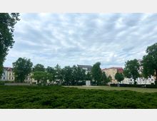 Grüne Parkfläche mit niedrigen Sträuchern im Vordergrund, umgeben von Bäumen und mehrstöckigen Wohngebäuden in hellen Farben im Hintergrund. Der Himmel ist überwiegend bewölkt, und ein kleiner, quadratischer Steinsockel steht auf einer Wiese in der Mitte.