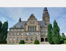 Historisches Gebäude aus Sandstein mit einem großen Uhrturm, steilem Schieferdach und zahlreichen Fenstern; davor gepflegte Grünflächen und hohe, schlanke Bäume.