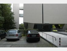 Mehrstöckiges modernes Gebäude mit geometrischer Architektur, breitem Gehweg mit Rampe im Vordergrund und einem kleinen Parkplatz mit zwei Autos links. Im Hintergrund sind Bäume und grüne Vegetation zu sehen.