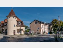 Straßenszene mit einem historischen Torbau namens "Potsdamer Tor", erkennbar an einem runden Turm mit spitzem Dach und Arkaden, flankiert von Wohnhäusern und einer Bahnschranke. Im Vordergrund verlaufen Kopfsteinpflaster und Gleise, umgeben von Verkehrsampeln und Straßenschildern.