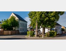 Straßenszene mit einem historischen Gebäude auf der linken Seite, beschriftet mit „Färberei-Reinigung“, sowie einem kleinen Platz mit einem Obelisken und einem großen Baum im Vordergrund. Auf der rechten Seite befinden sich weitere kleinere Gebäude, ein Parkplatz sowie Straßenschilder.