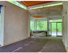 Ein leerer Innenraum in einem Gebäude, dessen Wände verputzt, aber noch nicht fertiggestellt sind. Große Fenster und Glastüren lassen viel Tageslicht herein und bieten einen Blick auf eine grüne Außenumgebung, der Boden ist mit dunkelgrauen Schutzmatten ausgelegt.