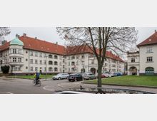 Wohnsiedlung mit mehreren mehrstöckigen Gebäuden im traditionellen Stil, erkennbar an roten Ziegeldächern und hellen Fassaden. Im Vordergrund eine Straße mit parkenden Autos, einem Fahrradfahrer und einem Baum, der teilweise die Sicht auf die Häuser verdeckt.