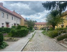 Eine kleine Wohnsiedlung mit Reihenhäusern, die beige und gelbe Fassaden sowie rote Ziegeldächer haben. Die Straße ist gepflastert, flankiert von Gehwegen, gepflegten Hecken, kleinen Gärten und vereinzelten Bäumen.