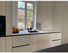 Moderne, helle Küche mit weißen Schränken und Arbeitsplatten aus schwarzem Material. Im Hintergrund ein großes Fenster mit Blick auf Bäume sowie eine Spüle und eine Obstschale.