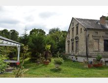 Ein älteres, teils verfallenes Wohngebäude mit verwitterter Fassade, umgeben von einem Garten mit Bäumen, Sträuchern und Blumen in Pflanzgefäßen. Links im Bild steht eine kleine, weiße Pergola mit einem Tisch darunter.