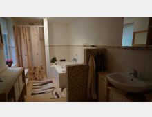 Helles Badezimmer mit Badewanne, Waschbecken und beigem Fliesenboden. Eine abgetrennte Dusche mit gemusterter Duschvorhang befindet sich links, daneben ein Fenster mit halbtransparenter Gardine; Handtücher hängen an einer gefliesten Wand.