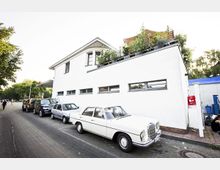 Ein modernes weißes Gebäude mit einem Flachdach und einem bepflanzten Balkon im oberen Bereich. Davor stehen mehrere geparkte Autos entlang einer gepflasterten Straße, umgeben von vereinzelten Bäumen und Fußgängern.
