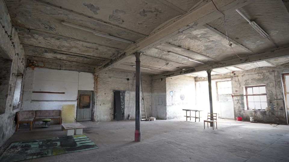 Großer, leerer Industrie- oder Lagerraum mit bröckelnder Decke, freiliegenden Trägern und Stützsäulen sowie alten Neonröhren. An den abgenutzten Wänden stehen vereinzelte Möbel und eine Leiter; Tageslicht fällt durch mehrere Fenster auf den Betonboden.