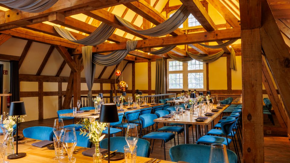 Festlich gedeckter Speisesaal in einem historischen Fachwerk-Dachgeschoss mit sichtbaren Holzbalken, schrägen Decken und drapierten Stoffbahnen unter dem Dach. Lange Tische mit Glaswaren und blau gepolsterten Stühlen stehen vor großen Sprossenfenstern und warmen Hängeleuchten.