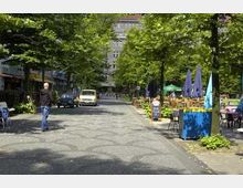 Straßenansicht mit Kopfsteinpflaster, gesäumt von Bäumen und Straßencafés mit Tischen und Schirmen. Im Hintergrund sind Wohn- oder Geschäftsgebäude sichtbar, während vereinzelt Fußgänger und ein geparktes Fahrzeug zu sehen sind.
