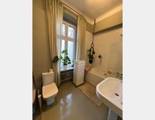 Helles Badezimmer mit einem großen Fenster, das von beigefarbenen Vorhängen eingerahmt ist. Das Zimmer verfügt über eine Toilette, ein Waschbecken, eine Badewanne mit Duschvorhanghalterung, eine Heizkörper unter dem Fenster sowie eine schmale, rollbare Schubladenkommode; Pflanzen dekorieren den Raum.