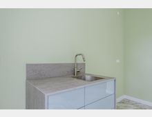 Moderne Küchenzeile mit grauer Beton-Arbeitsplatte und -Rückwand, integriertem Edelstahlspülbecken und hohem Wasserhahn. Glatte, hellgraue Fronten stehen vor einer hellgrünen Wand in einem schlichten Innenraum.