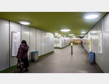 Unterirdischer Bahnhofsgang mit grauen Wänden und grünem Deckenbereich, beleuchtet von runden Deckenleuchten. An den Wänden befinden sich Fahrpläne, ein Fahrkartenautomat und Mülleimer, während im Hintergrund eine Wand mit bemaltem Blumenmotiv sichtbar ist.