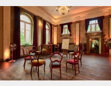 Eleganter Raum mit historischem Interieur, gekennzeichnet durch hohe Decken, große Fenster mit roten Vorhängen und eine dekorative Kronleuchterbeleuchtung. Im Raum stehen antike Stühle in einem Kreis, eine Staffelei mit Whiteboard sowie weitere Möbel wie ein Schreibtisch und ein Sofa.