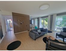 Wohnzimmer mit offener Gestaltung, hellen Wänden und großen Fensterfronten mit Vorhängen, die einen Blick auf einen Garten bieten. Die Einrichtung umfasst eine graue Couch mit Kissen, einen kleinen Holztisch und mehrere schwarze Teppiche auf hellem Holzboden; im Hintergrund ist ein Essbereich zu sehen.