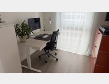 Helles, minimalistisches Homeoffice mit höhenverstellbarem Schreibtisch, Monitor, Schreibtischlampe und Drehstuhl auf grauem Holzfußboden. Ein großes Fenster mit transparentem Vorhang lässt Tageslicht herein; daneben steht ein rotes Schubladenregal.