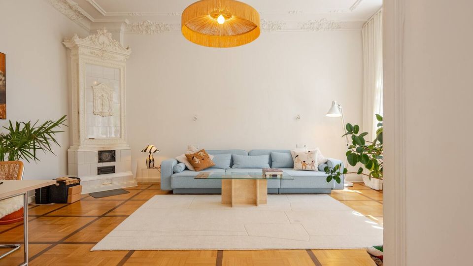 Helles Wohnzimmer mit hohen Decken, Stuckverzierungen und einem dekorativen weißen Kachelofen an der linken Wand. In der Mitte stehen ein hellblaues Sofa und ein Glastisch auf einem großen hellen Teppich; Parkettboden, eine große runde Deckenlampe, bodentiefe Vorhänge und mehrere Zimmerpflanzen rahmen den Raum.