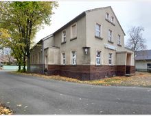 Ein zweistöckiges Gebäude mit hellem Putz, klassischen Fenstern und einer Fassade teils aus Ziegelsteinen. Links des Gebäudes stehen Bäume, einige mit gelbem Herbstlaub, und die Szenerie zeigt eine ruhige Straße in einem ländlichen Umfeld.