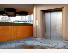 Außenbereich mit einer Aufzugsanlage in einem urbanen Umfeld. Die Wand ist mit orangefarbenen Fliesen verkleidet, und der Boden besteht aus Pflastersteinen und Beton. Im Hintergrund ist leicht ein Gebäude sowie Werbeschilder zu erkennen.