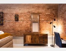 Schlafzimmer mit einer rustikalen Backsteinwand, einem Holzbuffet mit Spiegel, einer modernen Stehlampe, einem Schreibtisch mit schwarzem Stuhl und einem gepolsterten Bett im Vordergrund.