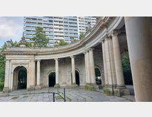 Historischer Pavillon mit runden Säulen und verzierten Bögen in einem urbanen Park, im Hintergrund ein modernes Hochhaus und grüne Bäume. Die Steinarchitektur zeigt Reliefs und ornamentale Details.
