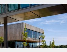 Ein modernes Gebäude mit einer Fassade aus Holzlatten und großen Glasfenstern, umgeben von jungen Bäumen und Laternen, unter einer transparenten Brücke in einem offenen, grünen Umfeld.