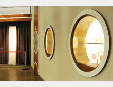 Gang in einem Gebäude mit hellen Wänden und runden Wandvitrinen, die durch Holzrahmen eingefasst sind und beleuchtete Exponate enthalten. Im Hintergrund ist ein mit dunklen Vorhängen abgetrennter Bereich und ein Ausstellungsschild zu sehen.