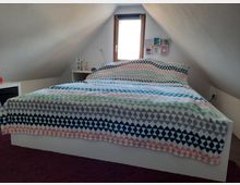 Ein Schlafzimmer in einem Dachgeschoss mit einer niedrigen, schrägen Decke. Im Zentrum steht ein Doppelbett mit einer gemusterten Decke, dahinter ein kleines Fenster und eine Wand mit dekorativen Bildern.