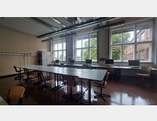 Ein Computerraum mit mehreren Arbeitsplätzen, bestehend aus Tischen, Stühlen und Monitoren, vor großen Fenstern, die viel Tageslicht hereinlassen. Im Hintergrund befinden sich ein weißer Schrank, eine Pinnwand und ein außen sichtbarer Schulhof mit einem Backsteingebäude und Bäumen.