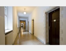 Ein langer, schmaler Flur mit weißen Wänden und hellbraunem Bodenbelag, gesäumt von dunklen Holztüren, die von hellem Fliesenmuster eingefasst sind. Links befinden sich Fenster mit einem Heizkörper darunter, und die Decke ist mit runden Hängelampen beleuchtet.