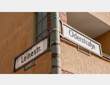 Straßenschild an einem graugrünen Metallpfosten mit den Bezeichnungen „Leinestr.“ und „Oderstraße“ vor der Fassade eines Gebäudes mit gelber Putzwand, roten Backsteinen und grünem Fensterrahmen.