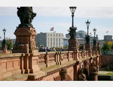 Rote Sandsteinbrücke mit dekorativen Balustraden und Statuen, flankiert von großen Laternen; im Hintergrund moderne Gebäude und die gläserne Kuppel des Berliner Reichstags, geschmückt mit deutschen Flaggen. Im Vordergrund ist eine Gruppe von Menschen auf der Brücke zu sehen.