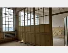 Ehemaliger Industrie- oder Werkstattraum mit großen Sprossenfenstern und einer hölzernen Trennwand mit Glaselementen. An der Wand sind ein Heizkörper und abblätternde Farbe zu sehen, was auf einen verfallenen Zustand hindeutet.