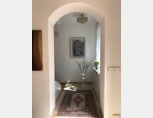 Ein schmaler, heller Flur mit einem gemusterten Teppich auf einem Fliesenboden, einem dekorativen Kronleuchter an der Decke und einem gerahmten Bild an der Wand. In der Mitte steht eine Vase mit künstlichen Blumen, rechts ist ein Fenster mit einem Radiator darunter, geschmückt mit einer Pflanze.