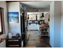 Eine moderne Küche mit weißer Ausstattung, dunkelbraunen Fliesen und hölzernen Elementen. Im Vordergrund ein kleiner Beistelltisch und eine Wand mit einem Bild eines verschneiten Gebirges. Im Hintergrund ist eine zentrale Arbeitsfläche mit Spüle, Schränken und Geräten sichtbar.