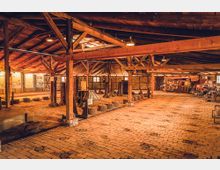 Großer Innenraum einer historischen Werkstatt oder Ausstellungshalle mit einer Holzbalkenkonstruktion und Backsteinboden. Im Raum sind Werkzeuge, Maschinen und Informationstafeln ausgestellt.