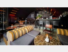 Eine moderne Bar mit dunkler, atmosphärischer Einrichtung, einschließlich einer großen Theke mit Barhockern, kleinen Marmortischen mit Kerzen und gepolsterten Sitzmöbeln in Streifenoptik. Im Hintergrund befinden sich weitere Sitzgelegenheiten und Regale mit Flaschen, beleuchtet von orangefarbenen Lichtstreifen an der Decke.