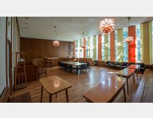 Ein mittelgroßer Saal mit Holzboden, großen vertikalen Fenstern mit farbigen Vorhängen in Gelb und Orange sowie modernen Kronleuchtern. Es gibt eine zentrale Bestuhlung mit Stühlen um Tische, eine erhöhte Holztribüne an der Rückwand und eine minimalistische Holzmöblierung.