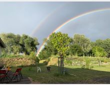 Ein Garten mit Sitzbereich und Holztisch im Vordergrund, umgeben von Rasen und Bäumen; ein Doppelregenbogen spannt sich über den bewölkten Himmel, während Sonnenlicht Teile der Landschaft beleuchtet.