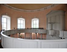 Innenansicht eines historischen Gebäudes mit halbkreisförmiger Empore aus weißem Holz und großen, bogenförmigen Fenstern an einer pastellfarbenen Wand. Rechts befindet sich eine erhöhte, dekorative Kanzel mit klassizistischen Elementen.