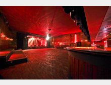Innenansicht eines Konzert- oder Clubraums mit Bühne im Hintergrund, roter Beleuchtung und Holzfußboden. Rechts im Bild ist eine geschwungene Bar sichtbar, links befinden sich erhöhte Sitzbereiche.