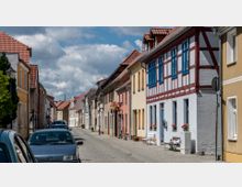Eine kleine, gepflasterte Straße mit geparkten Autos an der linken Seite, umgeben von zweigeschossigen Häusern mit bunten Fassaden und roten Ziegeldächern, darunter ein Fachwerkhaus mit blauen Fenstern. Einige Blumentöpfe und Sitzbänke stehen vor den Gebäuden, während der Himmel von dichten Wolken bedeckt ist.