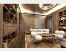 Luxuriöses Wohnzimmer mit hölzerner Wandverkleidung, Bücherregalen, einem hellen Chesterfield-Sofa, zwei Sesseln mit weißem Fellbezug, einem ovalen Couchtisch mit Metallgestell und einem großen Teppich mit orientalischem Muster. Eine gläserne Kronleuchter-Lampe hängt von der Decke.