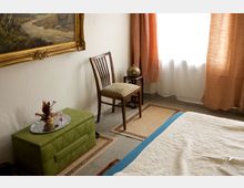 Ein schlicht eingerichtetes Zimmer mit einem Bett im Vordergrund, einem grünen Polsterhocker mit Tablett und Deko, einem Holzstuhl und einem kleinen Tisch mit Kupfervase daneben. Die Wand ist weiß, geschmückt mit einem gerahmten Landschaftsbild, und das Fenster ist mit orangen Vorhängen versehen.